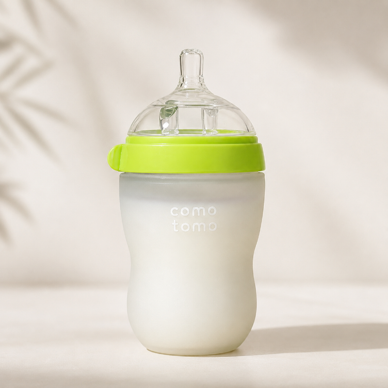 A baby bottle with a frosted white body, a bright lime green collar and loop, and a clear silicone nipple, shown on a warm beige background with soft leaf shadows.