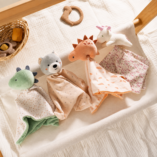 Three plush baby security blankets with animal and unicorn designs are arranged on a cream-colored textured blanket inside a wooden crib, alongside a woven baby toy basket.