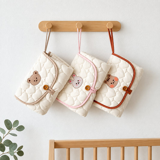 Three quilted baby pouches with cute animal faces hang from a wooden crib organizer bar on a white wall above a wooden crib. The pouches are cream, light pink, and terracotta, with matching trim and snap closures.