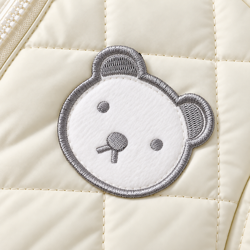 Close-up of a cream quilted puffer jacket featuring a white embroidered teddy bear face patch with gray stitching.