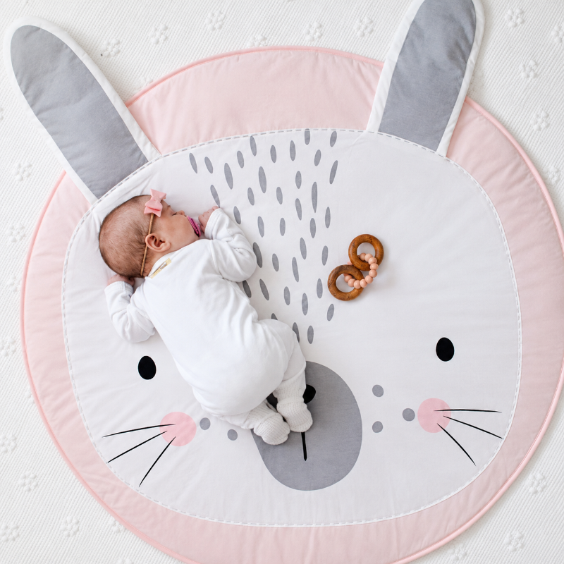 A baby in a white onesie lies on a round bunny-face play mat with gray ears, pink trim, and a wooden ring toy nearby, photographed from above on a white textured blanket.