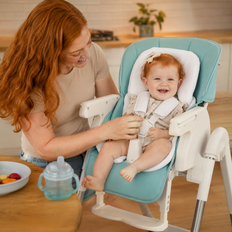 A baby with red hair sits in a high chair with a smiling woman adjusting the harness. The high chair is light teal and white, set in a warm kitchen environment with a wooden table and bowl of fruit in the foreground.