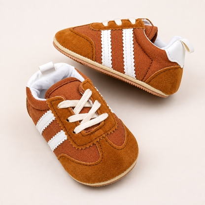 A pair of baby sneakers in rust brown suede and canvas with white stripes and white laces, displayed on a light beige background.