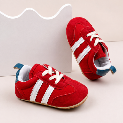 A pair of baby shoes in red suede with white scalloped stripes, white laces, and blue heel tabs, displayed on a light beige studio background with a white wavy box prop.
