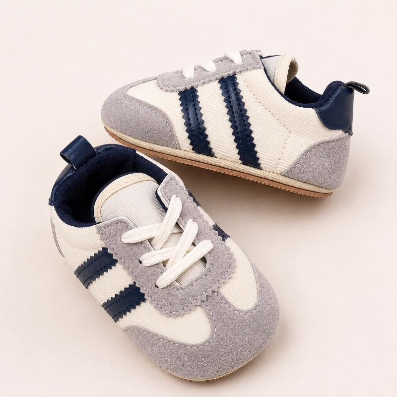 A pair of baby sneakers in cream, light gray, and navy blue, styled on a warm off-white background.