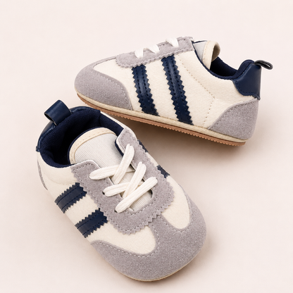 A pair of baby sneakers in cream, light gray, and navy blue, styled on a warm off-white background.