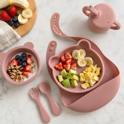 A coordinated children's breakfast set in dusty pink silicone, plate with a side tray, a bowl of yogurt with fruit and granola, a spoon and fork, and a small teapot, arranged on a light marble surface with fruit and a cloth visible.