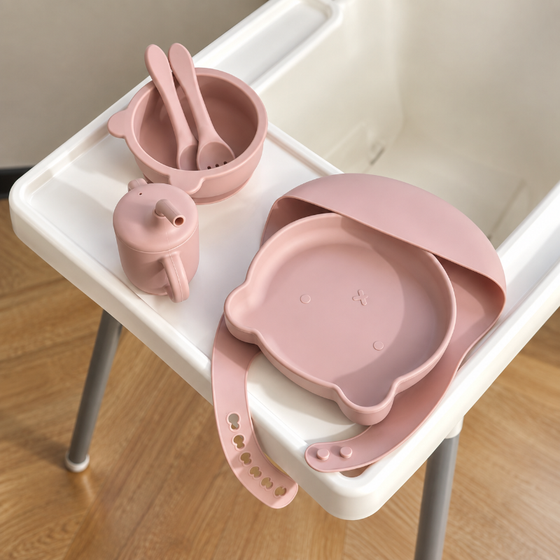 Pink silicone baby feeding set including a plate, bowl, spoon, small cup, and bib, arranged on a white high chair tray over a wooden floor.