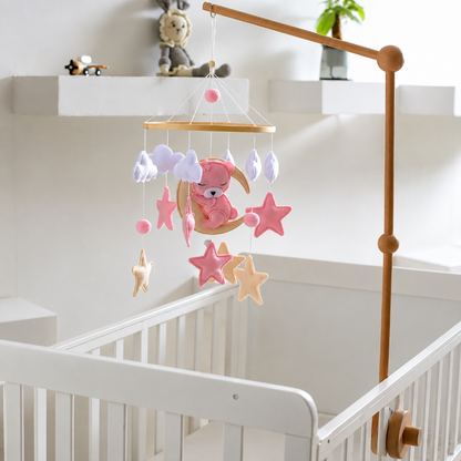 A wooden baby crib with a hanging mobile featuring a pink teddy bear on a crescent moon, pink and peach stars, and soft pastel cloud shapes, shown in a bright nursery setting.