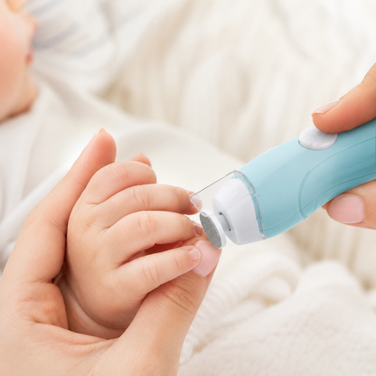 A close-up of a baby's hand being gently held while an adult uses a light blue and white electric nail grinder to trim the baby's fingernail. The background is a soft, warm white blanket.