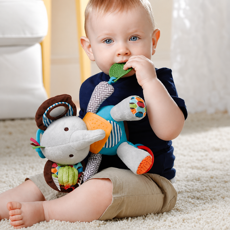 A baby sitting on a cream carpet holds and bites a colorful plush elephant toy. The baby is wearing a navy blue shirt and khaki shorts, with a soft white chair and curtain in the background.