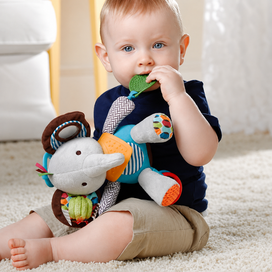 A baby sitting on a cream carpet holds and bites a colorful plush elephant toy. The baby is wearing a navy blue shirt and khaki shorts, with a soft white chair and curtain in the background.