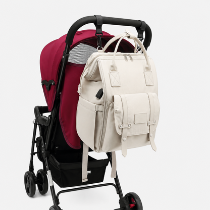 An off-white diaper backpack with multiple front pockets and top handles, shown hanging from the handlebar of a stroller with a burgundy liner, against a light gray background.