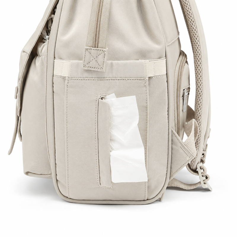 A light beige backpack shown from the back, featuring a large front pocket with a tissue dispenser slot holding a white tissue, side zippers, and padded shoulder straps on a clean white background.