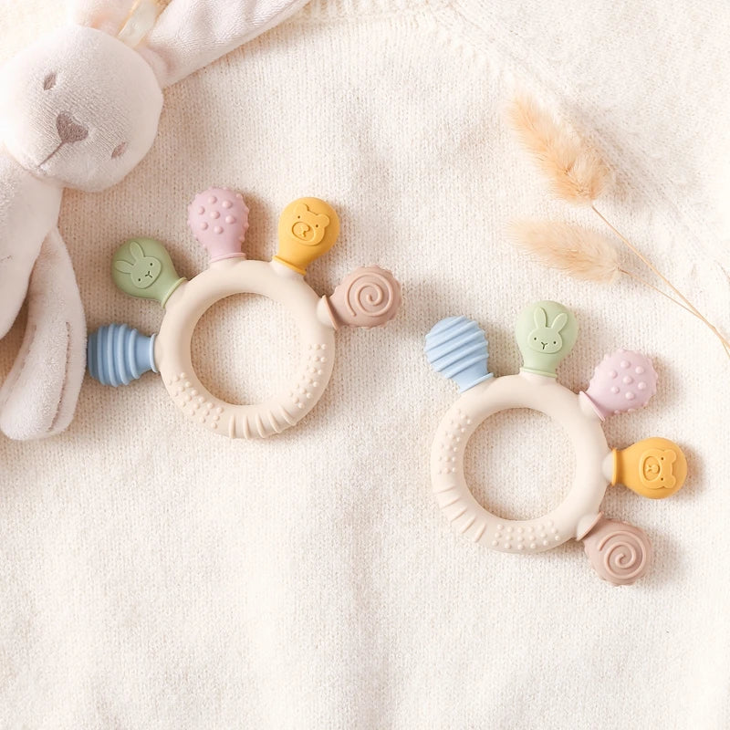 Two silicone baby teether rings with various colorful textures, designed to be held by little hands, placed next to a plush toy on a soft surface.