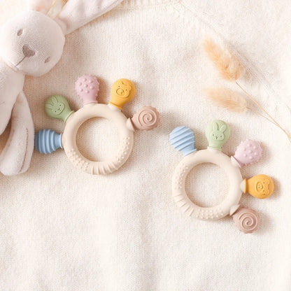Two silicone baby teether rings with various colorful textures, designed to be held by little hands, placed next to a plush toy on a soft surface.