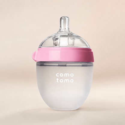 A baby bottle with a frosted white body, a pink collar, and a clear silicone top, featuring the 'como tomo' logo on the front, set against a warm beige background.