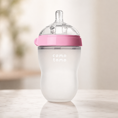 A baby bottle with a frosted white body, a pink collar, and a clear silicone nipple, standing on a light marble surface with a soft-focus warm background. The brand name 'como tomo' is printed on the front.