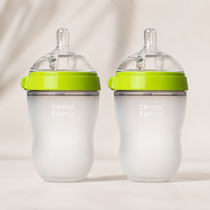 Two baby bottles with frosted white bodies, bright lime green bands, and clear silicone nipples, standing side by side on a light beige background. The bottles are branded with 'como tomo' in white text on the front.