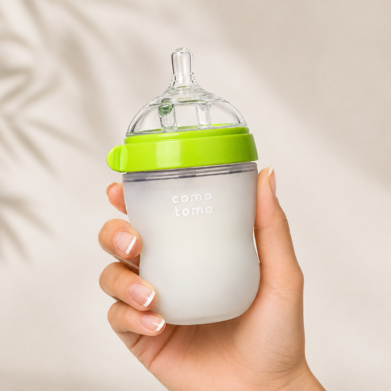 A hand holding a baby bottle with a frosted white body, a bright green collar, and a clear silicone nipple. The bottle has the brand name 'como tomo' printed on the front, set against a soft, light beige background with subtle leaf shadows.