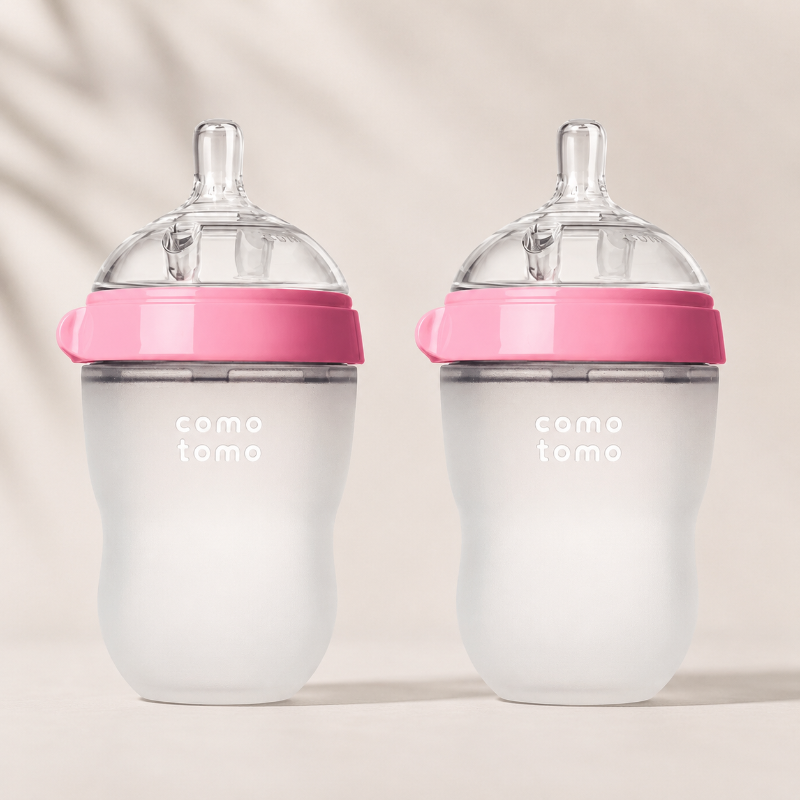 Two baby bottles with frosted white bodies, pink caps, and clear silicone nipples, displayed side by side on a light beige background with soft leaf shadows.