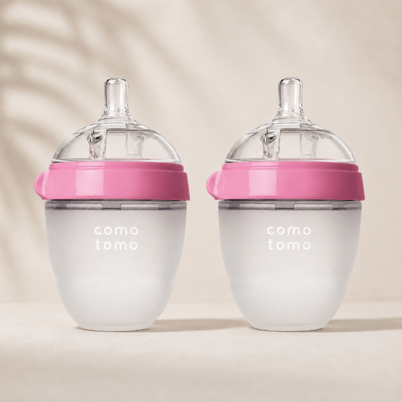 Two baby bottles with frosted white bodies, translucent caps, and bright pink bands, shown side by side on a warm beige surface with soft leaf shadows. The bottles have the text 'como tomo' printed on them.