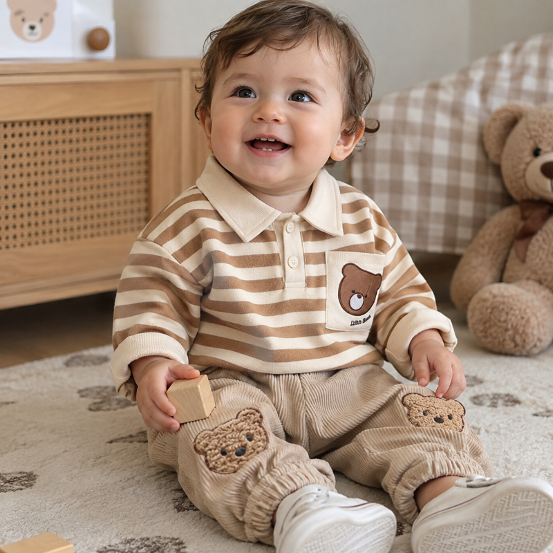 A baby sitting on a rug in a nursery, wearing a beige and brown striped long-sleeve polo shirt with a bear patch on the chest and matching corduroy pants with bear patches on the knees.
