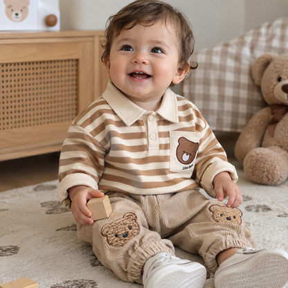 A baby sitting on a rug in a nursery, wearing a beige and brown striped long-sleeve polo shirt with a bear patch on the chest and matching corduroy pants with bear patches on the knees.