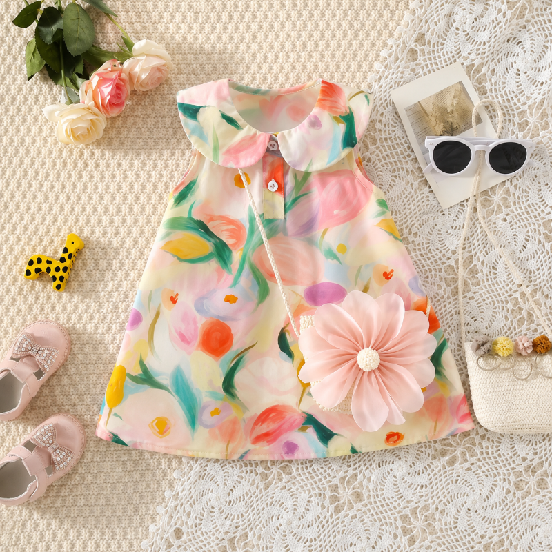 A child's sleeveless dress with a colorful floral print, styled in a flat lay with a pink fabric flower accessory, lace mat, sunglasses, shoes, and flowers.