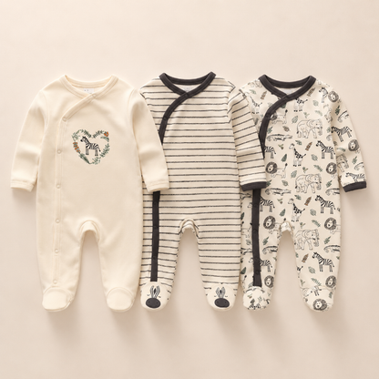 Three baby footed sleeper onesies laid flat on a light beige background. The left sleeper is cream with a floral heart design and the word 'Dus' inside.