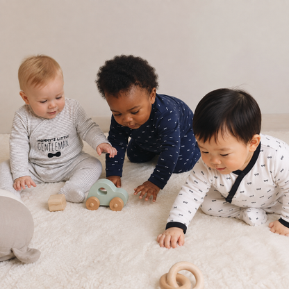 Three babies crawling and sitting on a white plush rug, wearing matching long-sleeve footless baby jumpsuits in gray, navy, and white with black accents. A wooden toy car and wooden teething rings are on the rug in front of them.