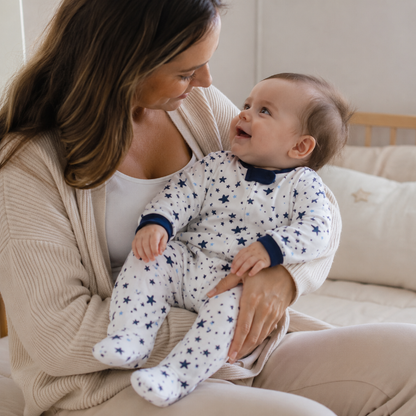 A baby wearing a white footed onesie with navy blue star patterns sits on a mother's lap. The mother is wearing a beige cardigan and cream pants, and they are sitting on a bed with neutral-colored pillows.