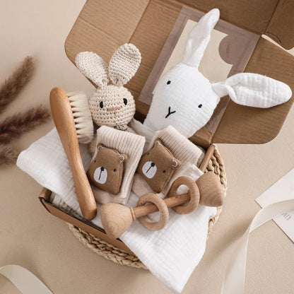 Baby care items including a rabbit plush toy, wooden brush, and rattle in a cardboard box on a beige surface.