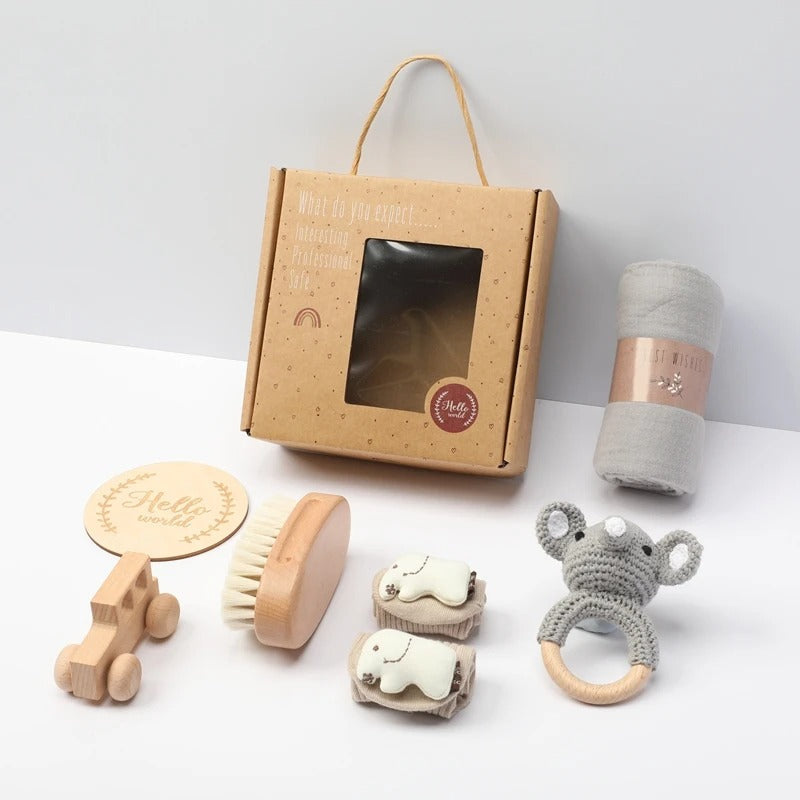 Baby gift set in a kraft paper box with a window, including a wooden toy car, brush, elephant teething rings, a knitted koala teether, and a rolled gray cloth, arranged on a white surface.
