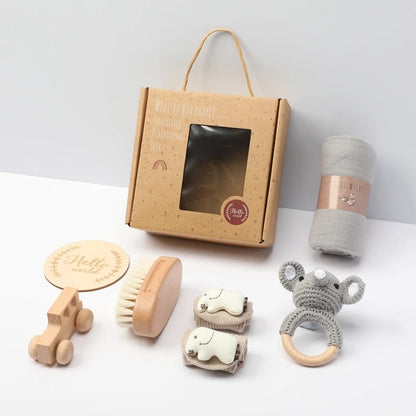 Baby gift set in a kraft paper box with a window, including a wooden toy car, brush, elephant teething rings, a knitted koala teether, and a rolled gray cloth, arranged on a white surface.