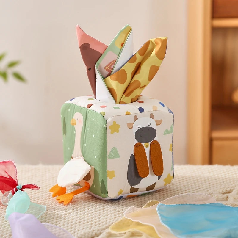 A colorful sensory tissue box designed for babies. The box is made from soft, safe materials with a red and blue face, and it contains several crinkly, multicolored scarves.