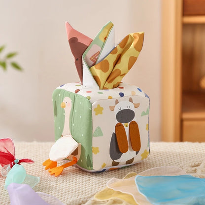 A colorful sensory tissue box designed for babies. The box is made from soft, safe materials with a red and blue face, and it contains several crinkly, multicolored scarves.