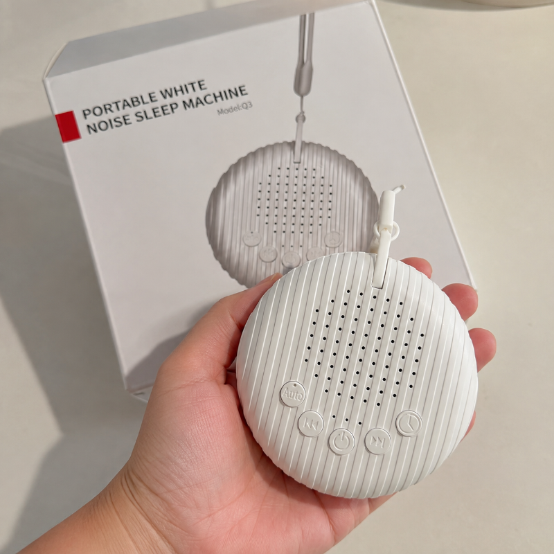 A hand holds a small round white portable noise sleep machine in front of its product box, which reads 'PORTABLE WHITE NOISE SLEEP MACHINE Model Q2'.